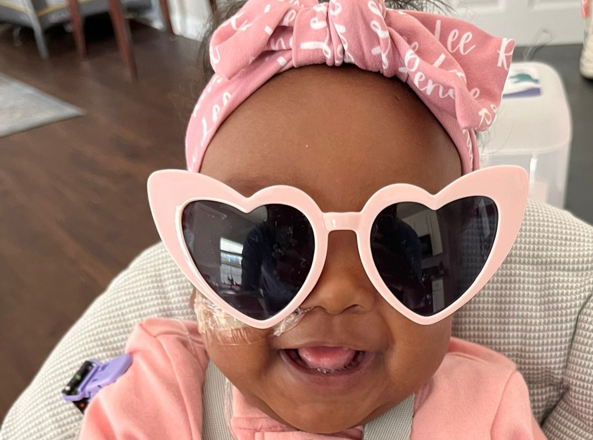 Baby Renee wearing pink heart sunglasses and pink headband, smiling big