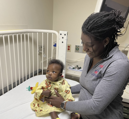 Mom smiling at baby Renee sitting in a yellow hospital gown on a hospital bed